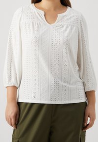 Biała bluzka z haftowanym wzorem oczek, o 1/4 rękawach i dekolcie w kształcie łezki. Miękka faktura i swobodny krój, noszona z oliwkowymi spodniami.
