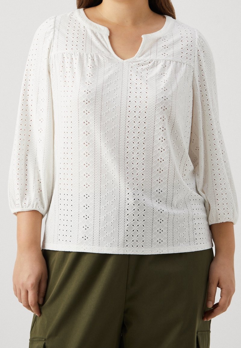 Biała bluzka z haftowanym wzorem oczek, o 1/4 rękawach i dekolcie w kształcie łezki. Miękka faktura i swobodny krój, noszona z oliwkowymi spodniami.