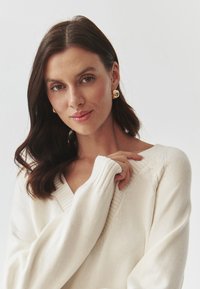 Pulover oversized de culoare crem, cu decolteu în V, accente din tricot cu nervuri și tricotaj texturat. Purtați cu cercei geometrice aurii.