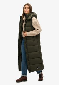 Langer, olivgrüner Puffer-Weste mit Kapuze, die gesteppte Abschnitte und seitliche Taschen aufweist. Getragen über einem beigen Strickpullover und Jeans, kombiniert mit braunen Stiefeln.