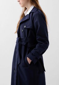 Trench coat bleumarin confecționat dintr-un material neted, cu butoane dublu rânduite, talie cu cordon și guler clasic.