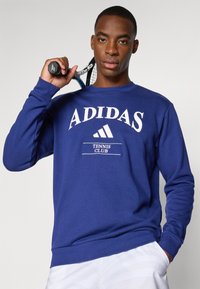 Blå Adidas sweatshirt med hvid tekst "ADIDAS" og "TENNIS CLUB." Blødt stof, lange ærmer, ribbede manchetter og kant. Tennisracket i hånden.