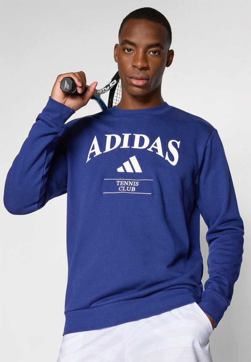 Sudadera azul de Adidas con el texto blanco "ADIDAS" y "TENNIS CLUB". Tejido suave, mangas largas, puños y dobladillo acanalados. Raqueta de tenis en la mano.