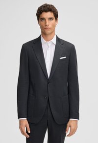 Giacca da uomo grigia scura in un tessuto testurizzato con revers a falda, due tasche frontali e un fazzoletto bianco, indossata sopra una camicia bianca.