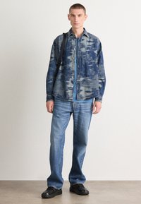 Blauwe denim shirt met abstract patroon, knoopsluiting aan de voorkant en borstzak, gecombineerd met een lichtblauw T-shirt, vervaagde blauwe jeans en zwarte schoenen.