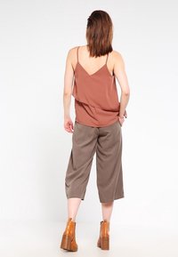 Top senza maniche marrone con design drappeggiato, abbinato a culotte beige larghe e stivaletti alla caviglia marroni con tacco. Tessuto dalla texture liscia.