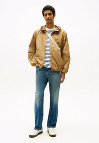 Rompevientos de color marrón claro con capucha, camiseta a rayas, vaqueros azules y zapatillas blancas. El rompevientos tiene una cremallera frontal y bolsillos laterales.