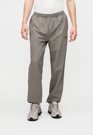 Άνδρας που φοράει γκρι παντελόνι με κορδόνι και ταιριαστά γκρι αθλητικά παπούτσια, στέκεται μπροστά σε ένα απλό λευκό φόντο.