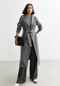 Manteau long en chevrons gris avec une taille ceinturée, associé à un pantalon large noir et des baskets blanches. Les accessoires comprennent un petit sac noir.
