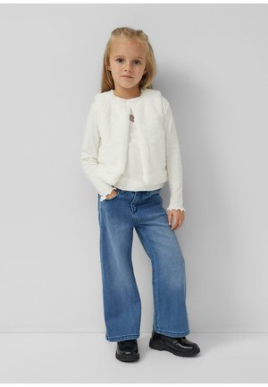 Dziewczyna stoi z ręką w kieszeni, ubrana w biały puszysty bezrękawnik, białą koszulę z długim rękawem, szerokie niebieskie dżinsy i czarne buty, na tle szarej ściany.