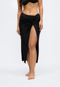Crna maksi suknja s detaljem uvijenog čvora u struku i dubokim prorezom iznad bedra, izrađena od glatkog, laganog materijala.