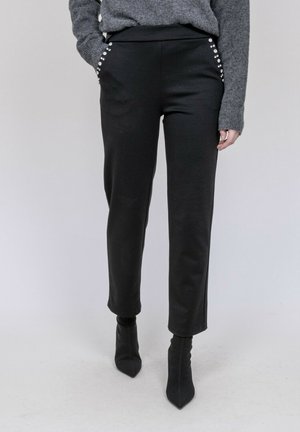 Personne portant un pantalon noir avec des poches ornées, un pull gris et des bottines noires pointues sur un fond neutre.