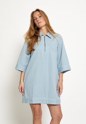 Frau mit langen blonden Haaren, die ein hellblau gestreiftes Kleid mit ellenbogenlangen Ärmeln und einem Reißverschluss vorne trägt.