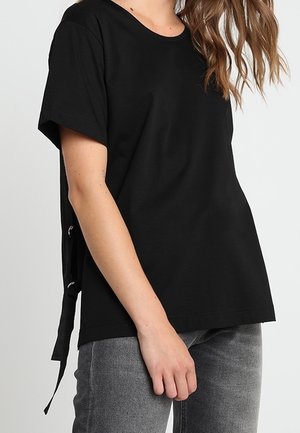 Black short-sleeve t-shirt made of cotton, featuring a rounded neckline and cinched lace-up detail at the back. Paired with grey jeans.