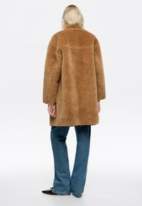 Abrigo de piel sintética marrón con diseño de cuello, forma oversized y mangas largas, combinado con unos vaqueros anchos azules y zapatos oscuros.