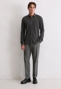 Młody mężczyzna stojący w ciemnoszarej koszuli na guziki, jasnoszarych dopasowanych spodniach i czarnych skórzanych butach, na tle jasnego, jednolitego tła.