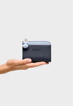 Dłoń trzymająca mały, czarno-niebieski portfel TOUS z zawieszką na suwaku w kształcie misia na jednolitym tle.