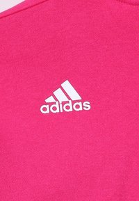 Rosa sporttröja med en vit Adidas-logga på framsidan. Materialet verkar mjukt med en strukturerad yta. Rund halsdesign.