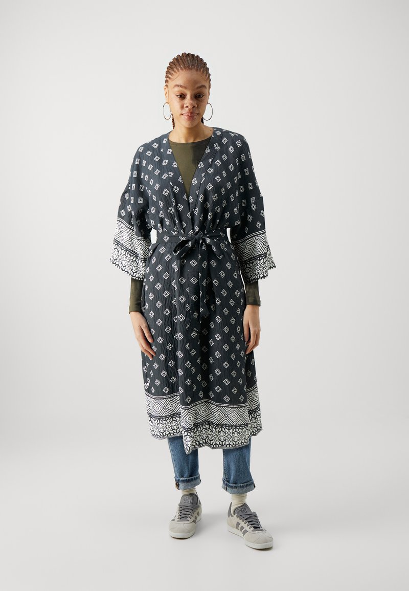 Dlhý čierny kimono so vzorom bielych diamantov, s tromi �štvrtinovými rukávmi a krajkovým lemom. Nosené cez zelený top a modré džínsy so teniskami.