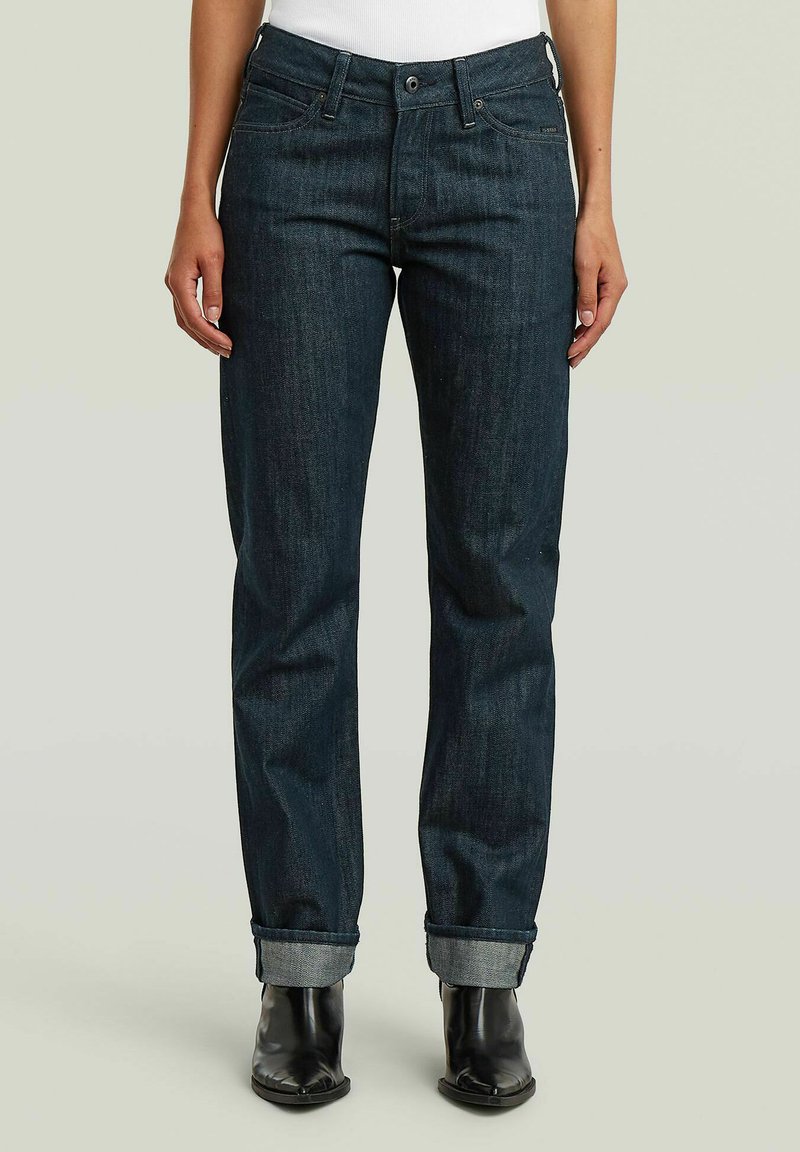 Person trägt dunkelblaue, aufgekrempelte Jeans, weißes Top und schwarze Stiefeletten, steht vor einem schlichten hellen Hintergrund.