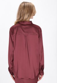 Bordó szatén ing hosszú ujjakkal, hátul redővel és hegyes gallérral, gombos mandzsettákkal és sima, fényes felülettel.