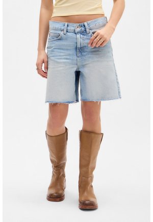 Vrouw met lichtblauwe spijkershorts met rafelige zomen en bruine leren laarzen tot halverwege de kuit, staand tegen een witte achtergrond.