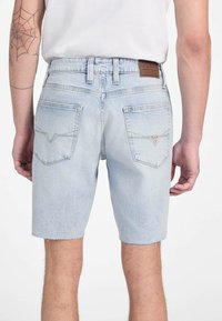 Shorts en denim bleu clair avec un ourlet effiloché, cinq poches et un patch en cuir au dos de la taille. Présente une texture décolorée.