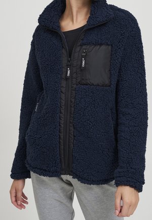 Marineblaue Fleecejacke mit Struktur, schwarzer gesteppter Brusttasche und Reißverschluss, getragen über einem schwarzen Oberteil und kombiniert mit hellgrauen Hosen.