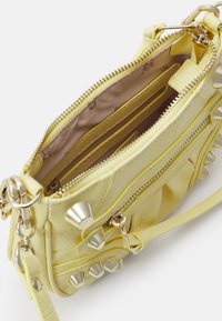Petit sac à main jaune avec des clous argentés, une fermeture éclair sur le dessus et un intérieur ouvert montrant une doublure en tissu et des poches intérieures avec une sangle de poignet attachée.