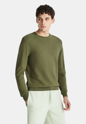 Młody mężczyzna w oliwkowozielonym swetrze z okrągłym dekoltem i jasnobeżowych spodniach, stoi z rozluźnionymi rękami, patrząc nieco na bok.