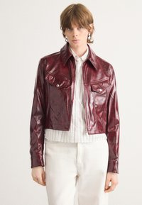 Burgundy faux leather cropped jacket with a classic collar, two chest pockets, and silver hardware details. Smooth texture with subtle sheen.