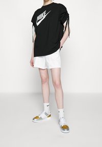 Musta ylipitkä Nike t-paita, jossa on suuri valkoinen logo, yhdistettynä valkoisiin shortseihin ja hopea/kultaisiin lenkkikenkiin. Valkoiset sukat, joissa on logokuvioita.