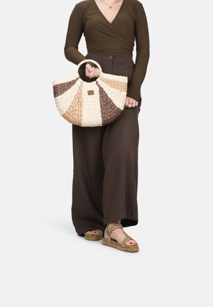 Borsa a mezzaluna intrecciata con strisce marroni, crema e beige; presenta un manico circolare e un piccolo logo quadrato. Il modello indossa pantaloni a gamba larga marroni.