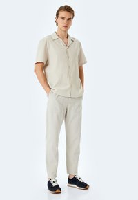 Camicia beige a maniche corte con bottoni, realizzata in un tessuto testurizzato, abbinata a pantaloni grigi chiari dal taglio morbido e sneakers blu navale con lacci bianchi.