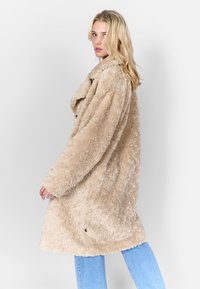 Faux fur jas in beige met een losse pasvorm, grote kraag en zichtbare knopen. Zachte, gestructureerde oppervlak, gecombineerd met lichtblauwe jeans.