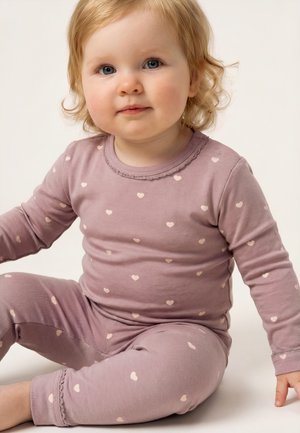 Småbarn med lyst krøllet hår, der sidder, iført mauve pyjamas dekoreret med små hjerte mønstre på en ensfarvet baggrund.