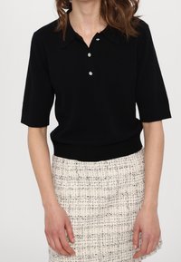 Polo suéter de manga corta negro con cuello, con tres botones decorativos. Combinado con una falda texturizada de color claro.