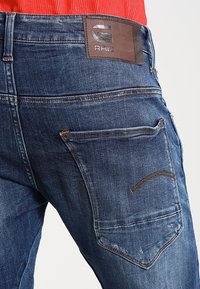 Tummansiniset farkut, joissa on nahkainen logo-ompelu, oranssia ompelua ja klassinen viisi-taskuinen muotoilu. hieman kuluneen näköinen pinta.