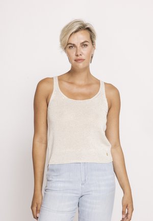 Blonde Frau mit kurzen Haaren, die ein beiges, ärmelloses Stricktop und hellblaue, hoch taillierte Jeans trägt und vor einem schlichten weißen Hintergrund steht.