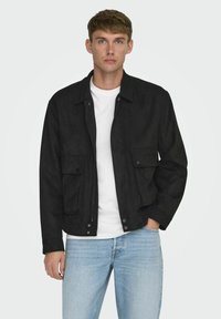 Svart mockajacka med två framfickor, tryckknappar och dragkedja framtill. Bärs över en vit t-shirt och ljusblå jeans.