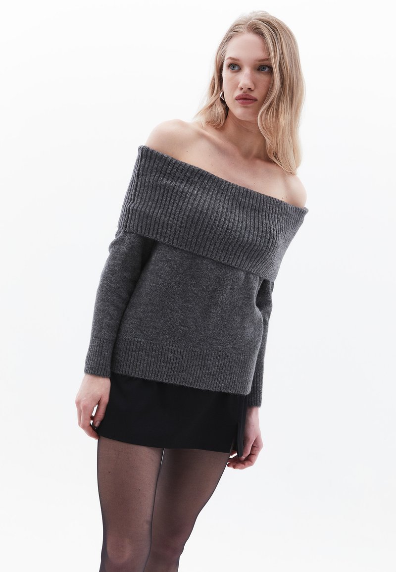 Ciemnoszary sweter typu off-the-shoulder z prążkowanymi detalami, długimi rękawami i miękką fakturą, w połączeniu z czarną spódnicą i transparentnymi rajstopami.