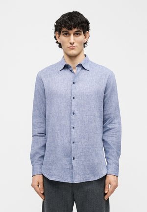 Jeune homme aux cheveux bouclés portant une chemise bleu clair à boutons et un pantalon gris foncé, debout devant un fond blanc uni.