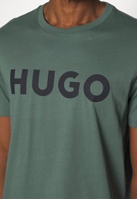 Zelené bavlněné tričko s kulatým výstřihem a krátkými rukávy má na hrudi velké černé logo "HUGO". Hladká textura.