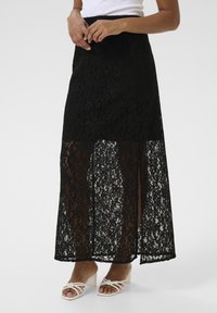 Jupe maxi en dentelle noire avec un motif floral, des sections transparentes et une taille ajustée. Complétée par des talons blancs à lanières.