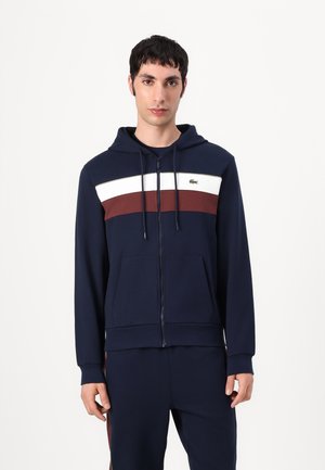 Marinblå hoodtröja med dragkedja som har vita och bourgognerandsömmen horisontellt över bröstet, snören och framfickor. Lacoste-logotyp på bröstet.