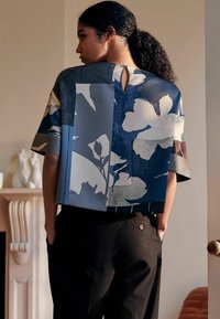Kortærmet top med blomsterprint i nuancer af blå og hvid, med en åbning i ryggen og en afslappet pasform, parret med sorte bukser.
