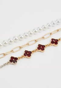 Three bracelets: white pearls, gold chain links, and gold with red gemstone flower-shaped charms on a white background.