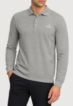 Mann trägt ein graues langärmeliges Polohemd mit drei Knöpfen und schwarze Hose, Hände entspannt seitlich, schaut nach vorne.