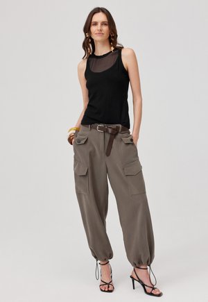 Kobieta stoi, ma na sobie czarny bezrękawnik z przejrzystym dekoltem, luźne khaki spodnie cargo z paskiem i czarne sandały na wysokim obcasie z paskami.