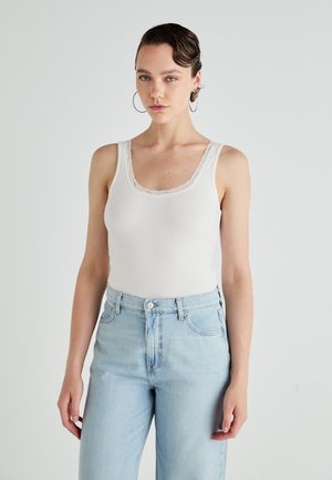Débardeur blanc à côtes avec un bord en dentelle au niveau de l'encolure dégagée, associé à un jean taille haute bleu clair avec des poches classiques.